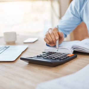 Close up detail of professional serious accountant sitting in light office, checking company finance profits on calculator, writing down results in notebook. Business concept